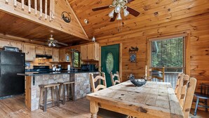 A wooden cabin interior with a dining table, chairs, a kitchen area, and a ceiling fan.