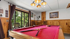 A pool table with a red felt surface, wooden surroundings, and a window with curtains.