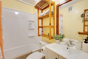 Een badkamer met een wit bad, een toilet, een wastafel met spiegel en een plank met handdoeken en toiletartikelen.