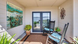 A covered porch with a view of the ocean, a rocking chair, a potted plant, and a large abstract painting. - Puget Sound Views: Des Moines Apt w/ Patio (Des Moines)