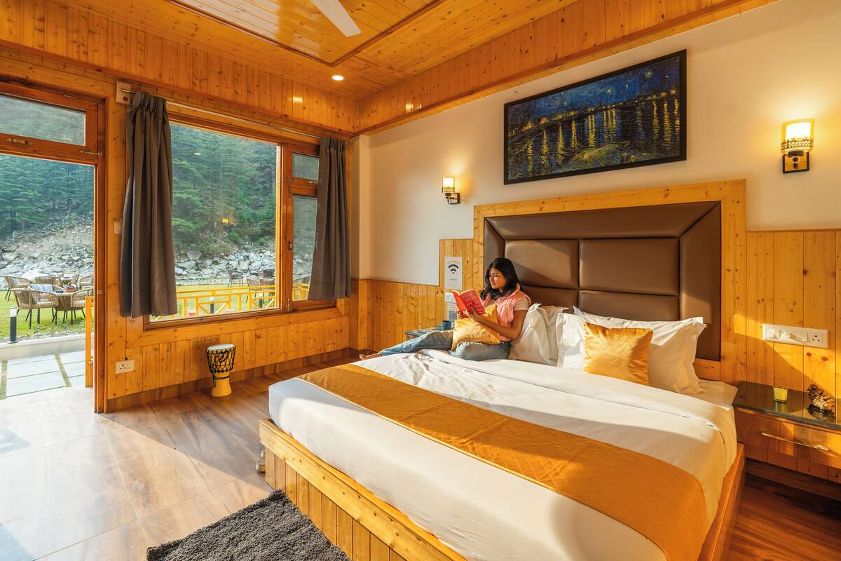 A wooden-furnished bedroom with a large bed, a painting above the bed, and a view of a rocky landscape through the window.