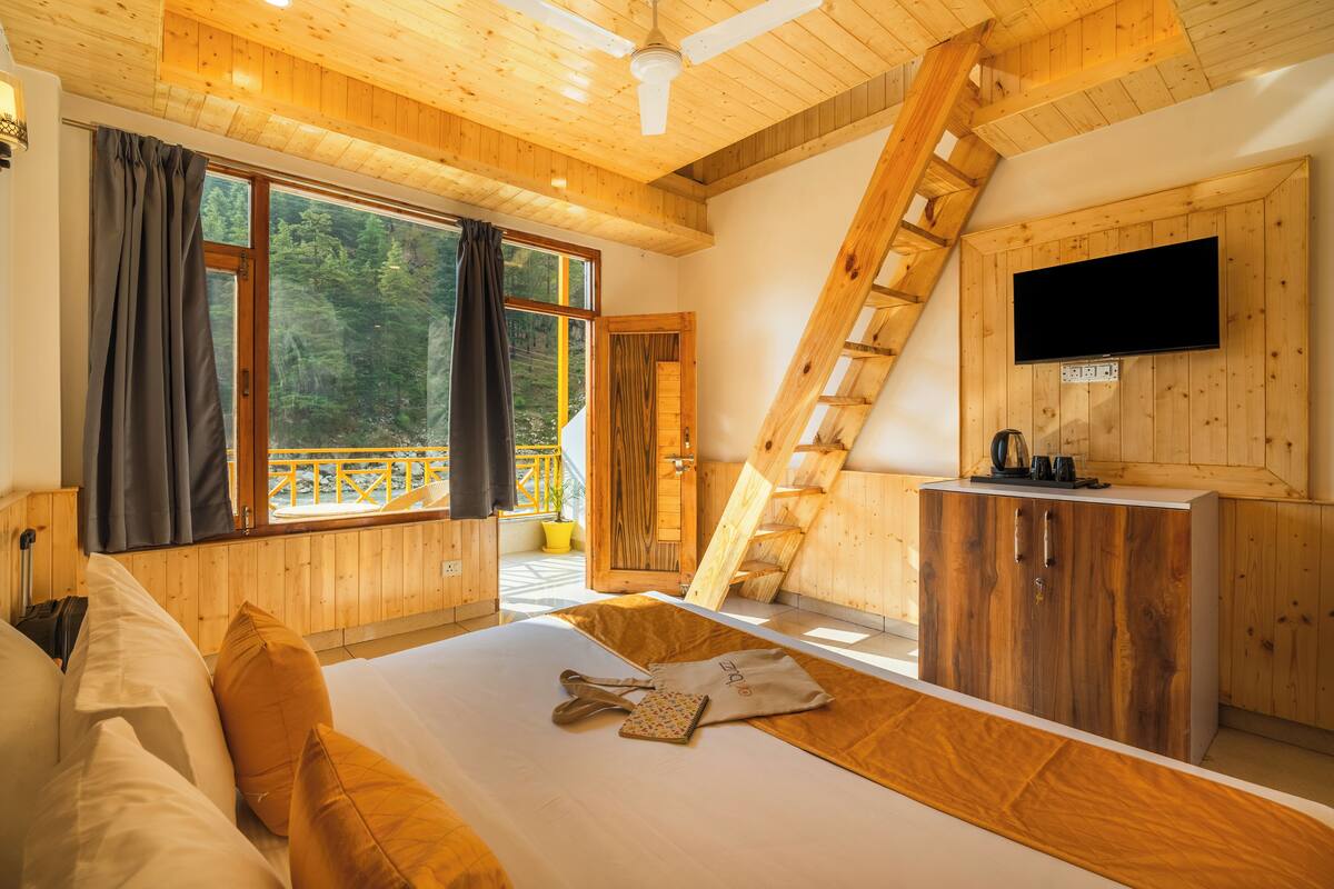 A wooden cabin bedroom with a bed, a TV mounted on the wall, and a large window overlooking a forest.