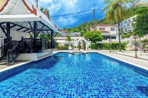 A swimming pool with a tiled deck, a covered seating area, and a view of residential buildings in the background. - Villa Atchara (Patong)