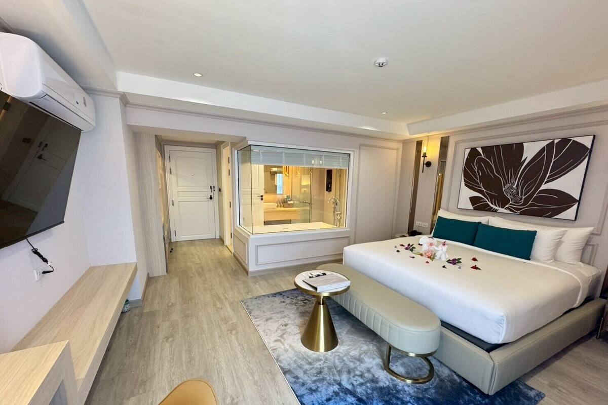 Una habitación de hotel moderna con una cama grande, una zona de estar, una mesita y un baño visible a través de una puerta de cristal.