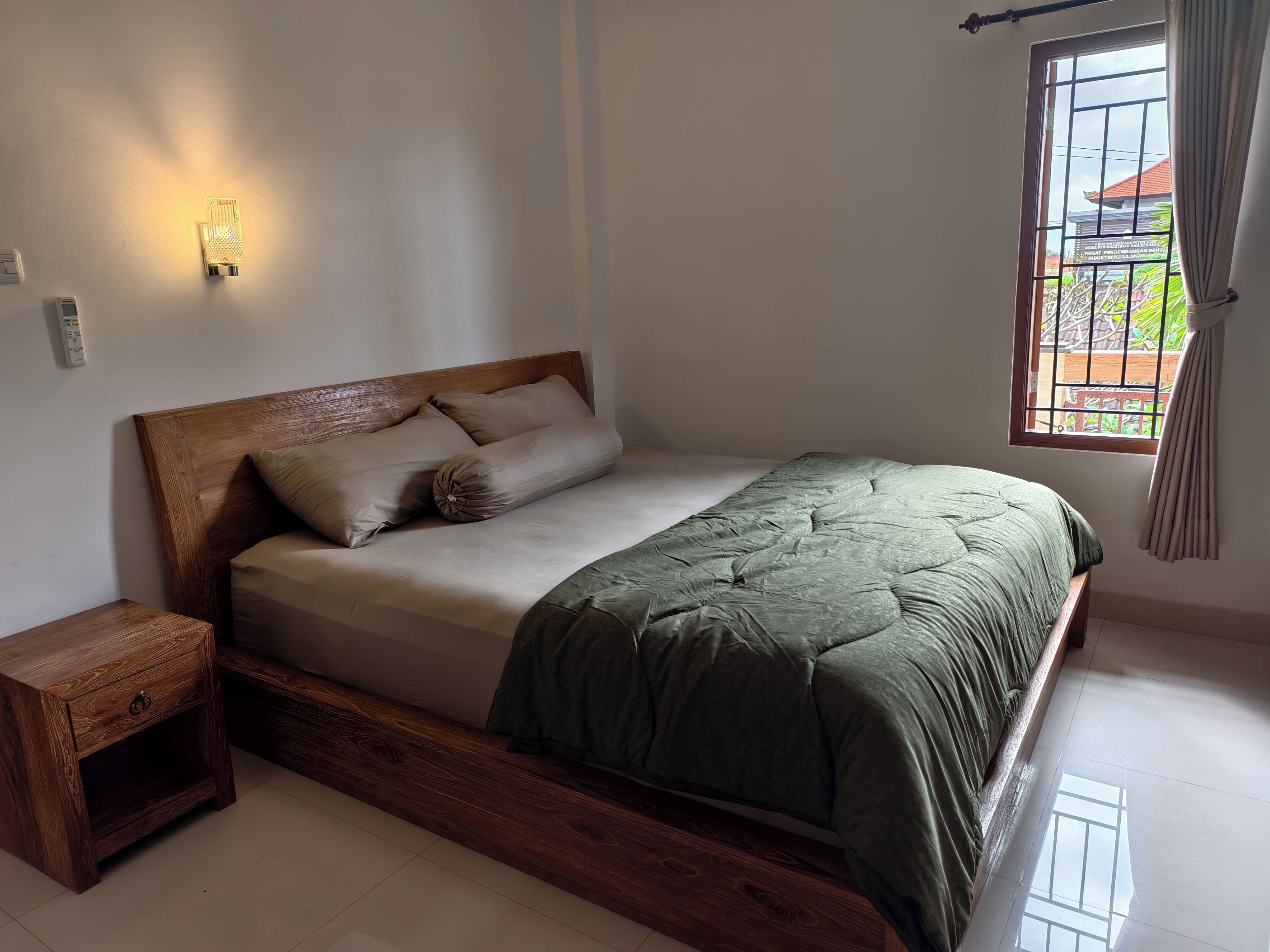 Un dormitorio con cabecera de madera, colcha verde, mesita de noche y ventana con cortinas.