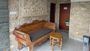 A room with a carved wall, a wooden sofa, a small stool, and a kitchen area. - Ayuna Guest House (Celuk)