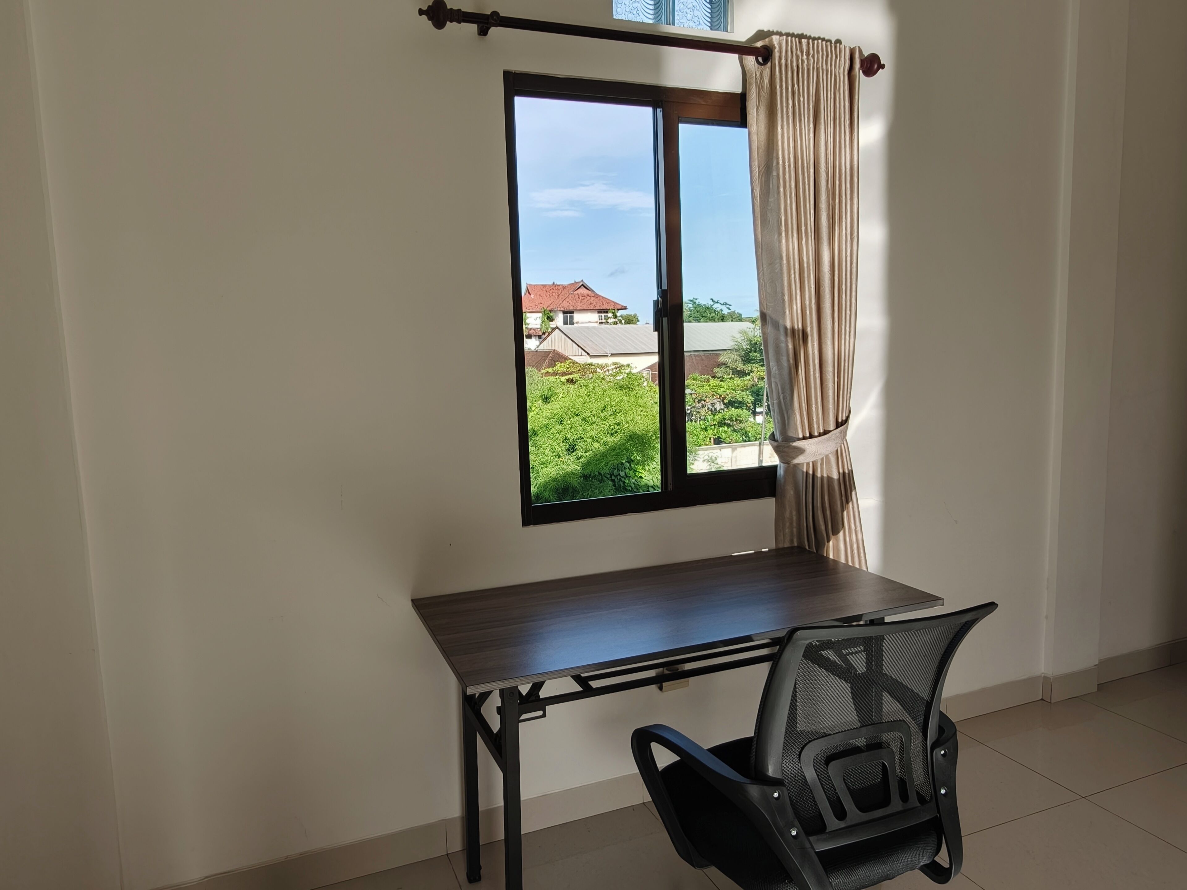 A room with a desk and chair, a window with curtains, and a view of greenery and buildings outside.