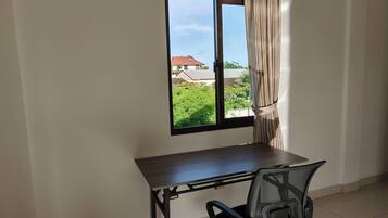 A room with a desk and chair, a window with curtains, and a view of greenery and buildings outside.