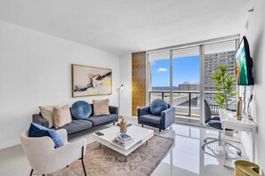 A modern living room with a gray sofa, a white coffee table, and a gray armchair. A large window offers a view of the city.