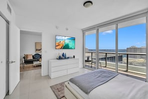 A modern bedroom with a large bed, a TV on the wall, a balcony with a city view, and a separate living area with a sofa and coffee table.