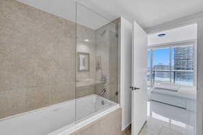 A modern bathroom with a glass-enclosed shower, a bathtub, and a view of a cityscape through a large window.