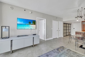 A modern living room with a flat-screen TV, a white console table, a dining area with a glass table and chairs, and a hallway with white doors.