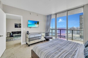 Un dormitorio con una cama grande, televisión, cómoda de cristal y vistas a la ciudad.