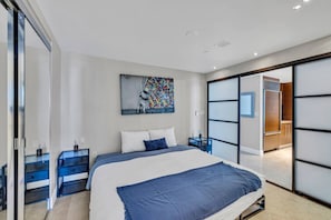 Un dormitorio moderno con una cama grande, un separador de vidrio, un escritorio pequeño y un cuadro abstracto de colores.