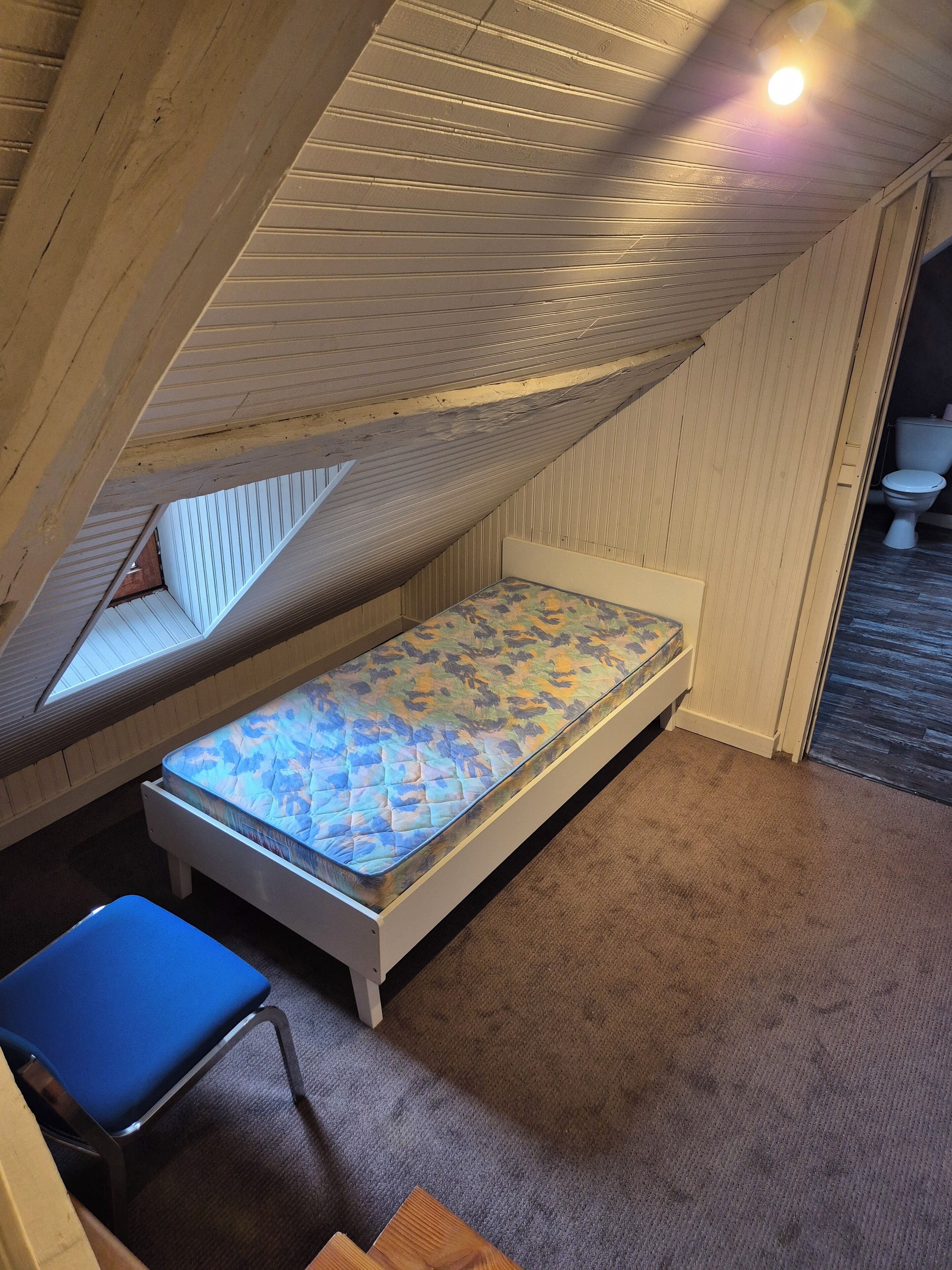 Ett litet rum med ett sluttande tak, en säng, en stol och en toalett som syns i det intilliggande rummet.