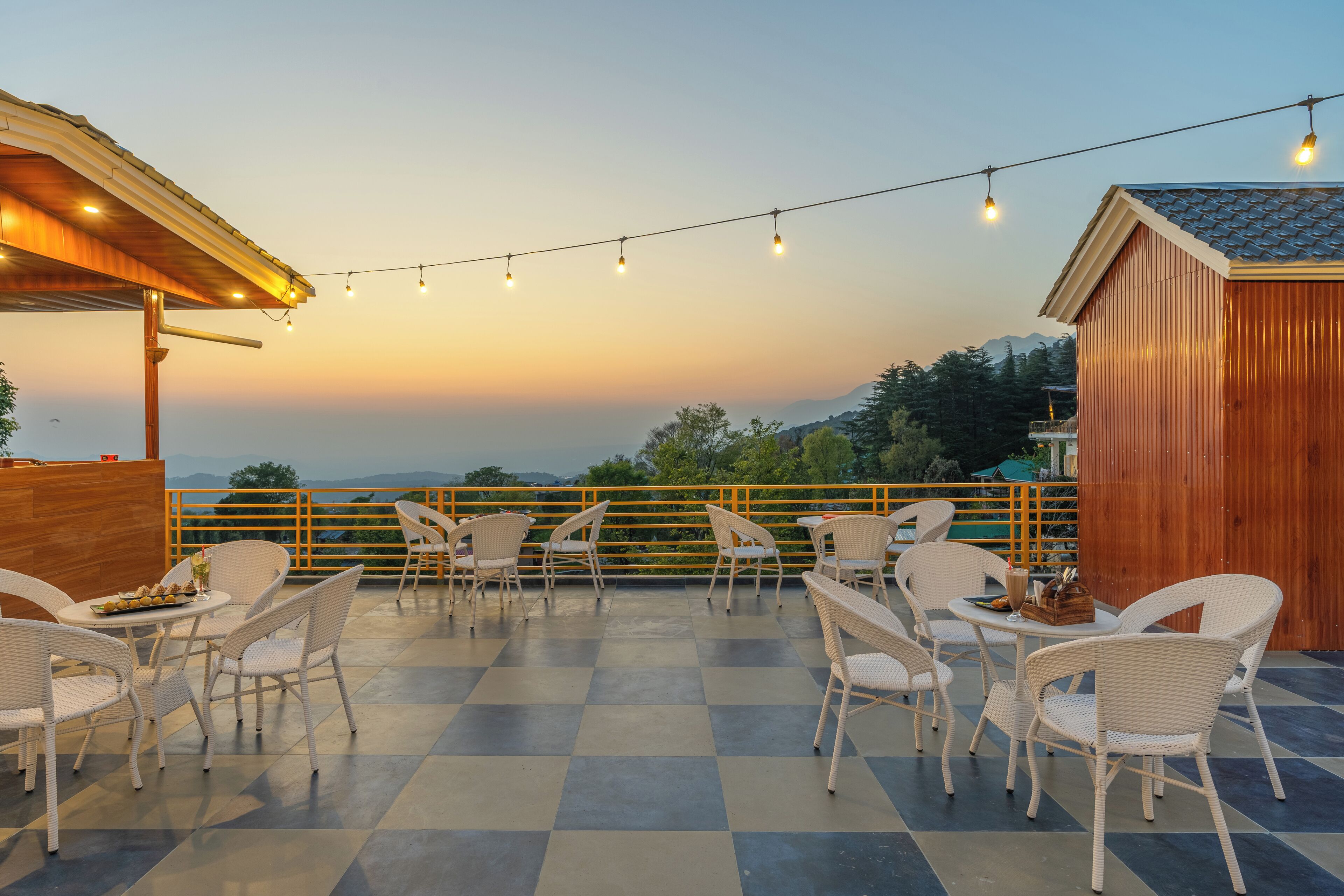 A terrace with checkered flooring, white wicker chairs, and tables, overlooking a scenic view with mountains in the distance.
