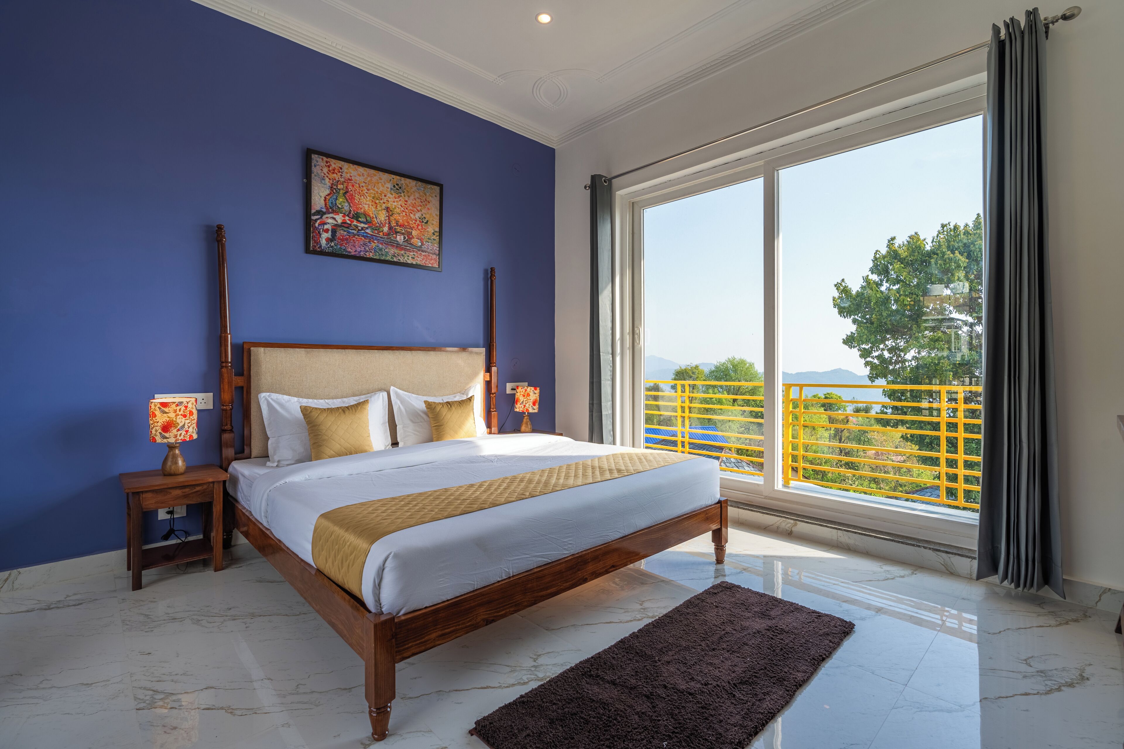A bedroom with a large bed, a nightstand with a lamp, a painting on the wall, and a view of a balcony with yellow railings.