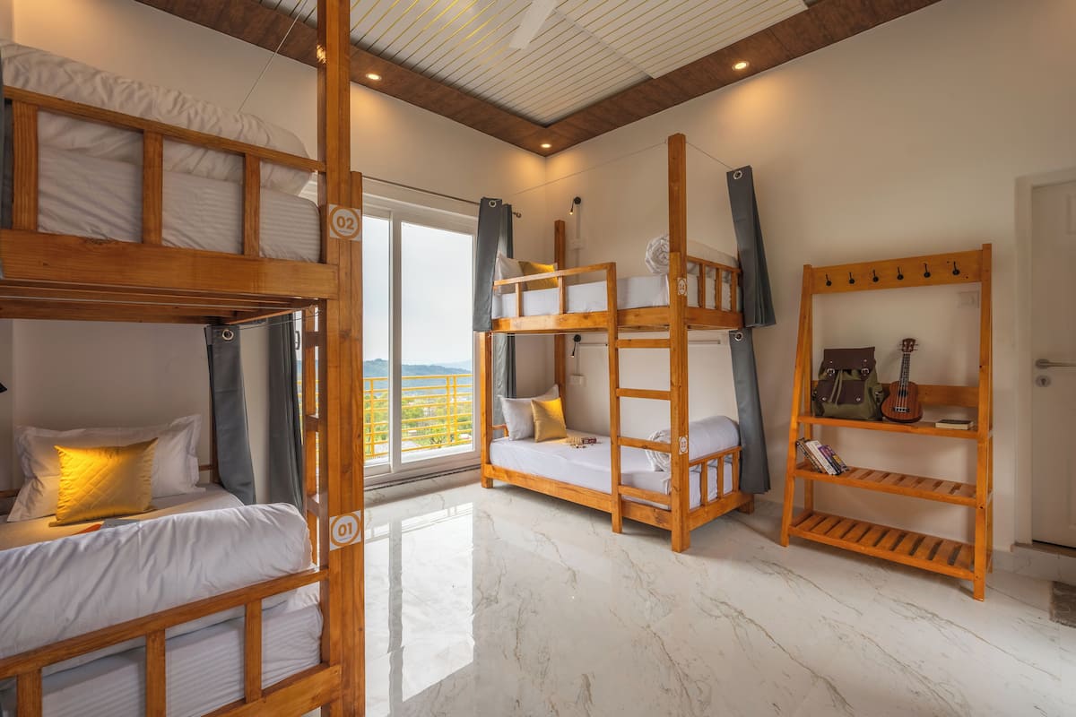 A room with two bunk beds, a wooden shelf with a guitar, and a view of a green landscape through a large window.