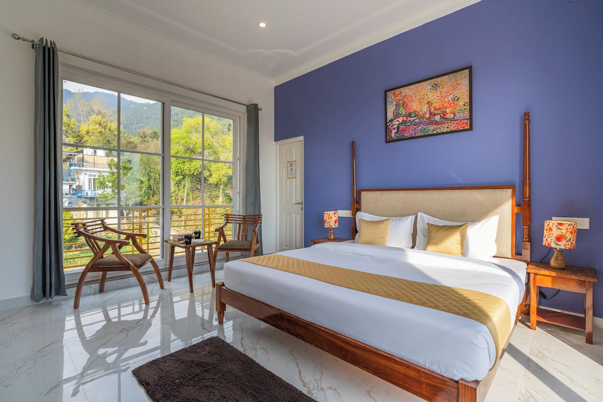 A bedroom with a large bed, two bedside tables with lamps, a view of greenery through a window, and a painting on the wall.