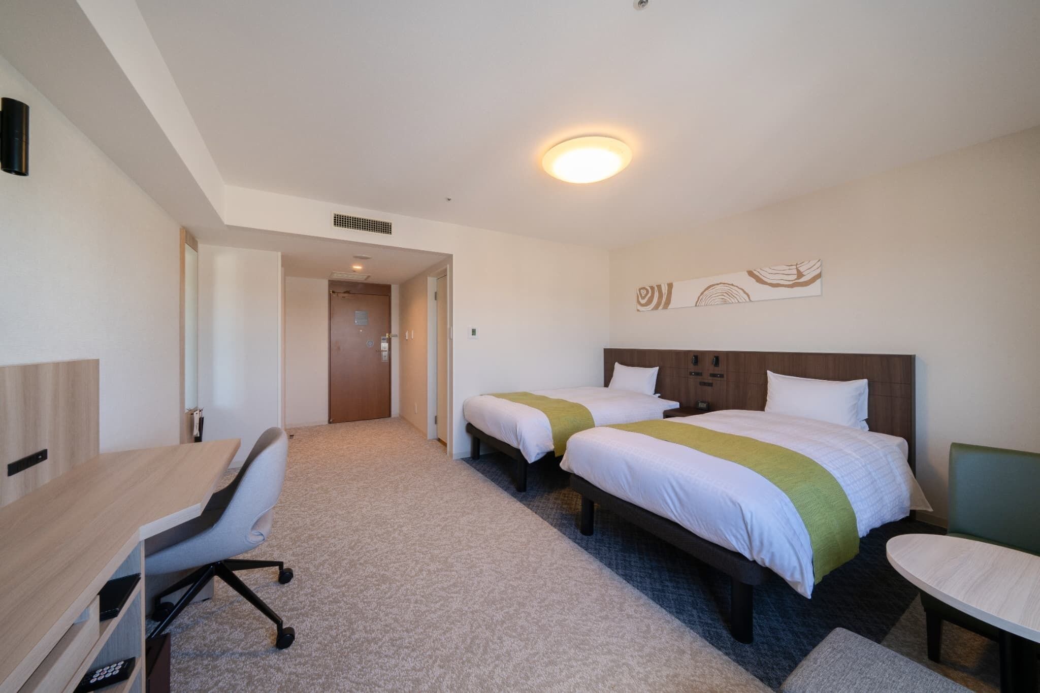 A hotel room with two beds, a desk, and a chair. There is a door leading to another room.