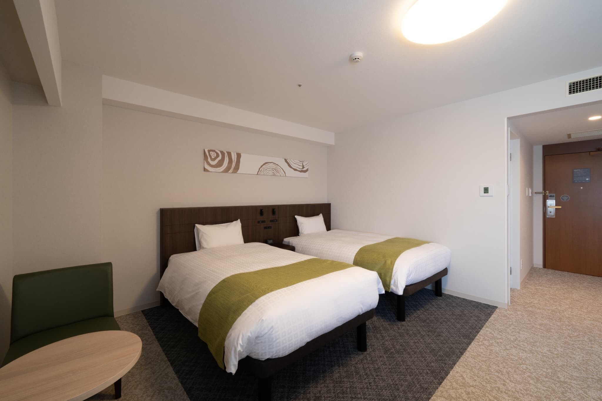 A hotel room with two beds, a green chair, a small table, and a wall-mounted artwork.