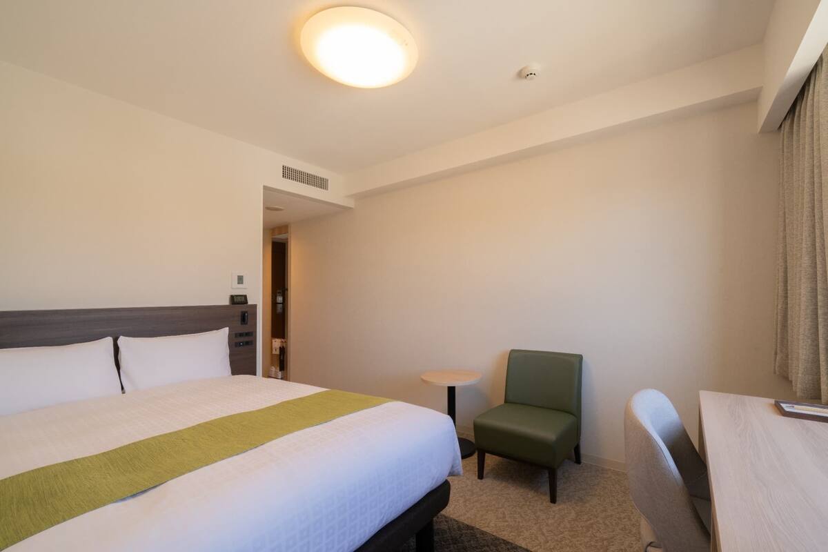 Una habitación de hotel con una cama grande, una mesita pequeña, una silla verde y un escritorio.