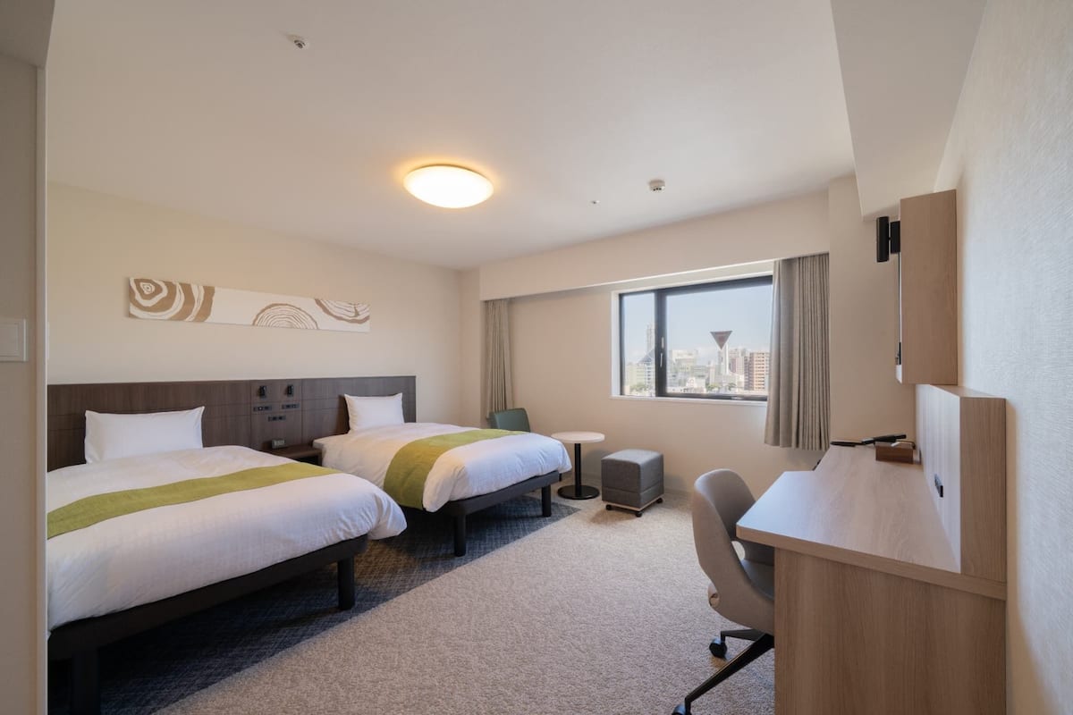 Habitación de hotel con dos camas, un escritorio, una silla, una mesita y una ventana con vista a la ciudad.