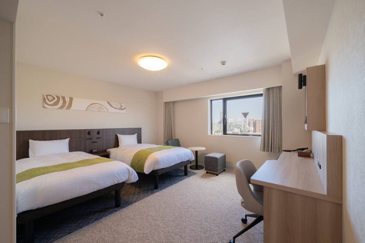 Habitación de hotel con dos camas, un escritorio, una silla, una mesita y una ventana con vista a la ciudad.