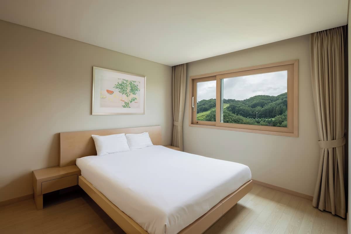 A neatly made bed in a room with a large window offering a view of a green landscape.