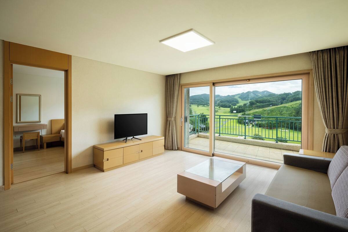 A modern living room with a flat-screen TV, a sofa, a coffee table, and a sliding glass door leading to a balcony with a view of green hills.