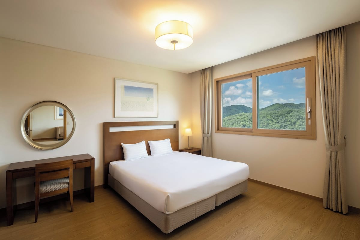 A hotel room with a large bed, a desk, a chair, a round mirror, a framed picture, and a window with a mountain view.