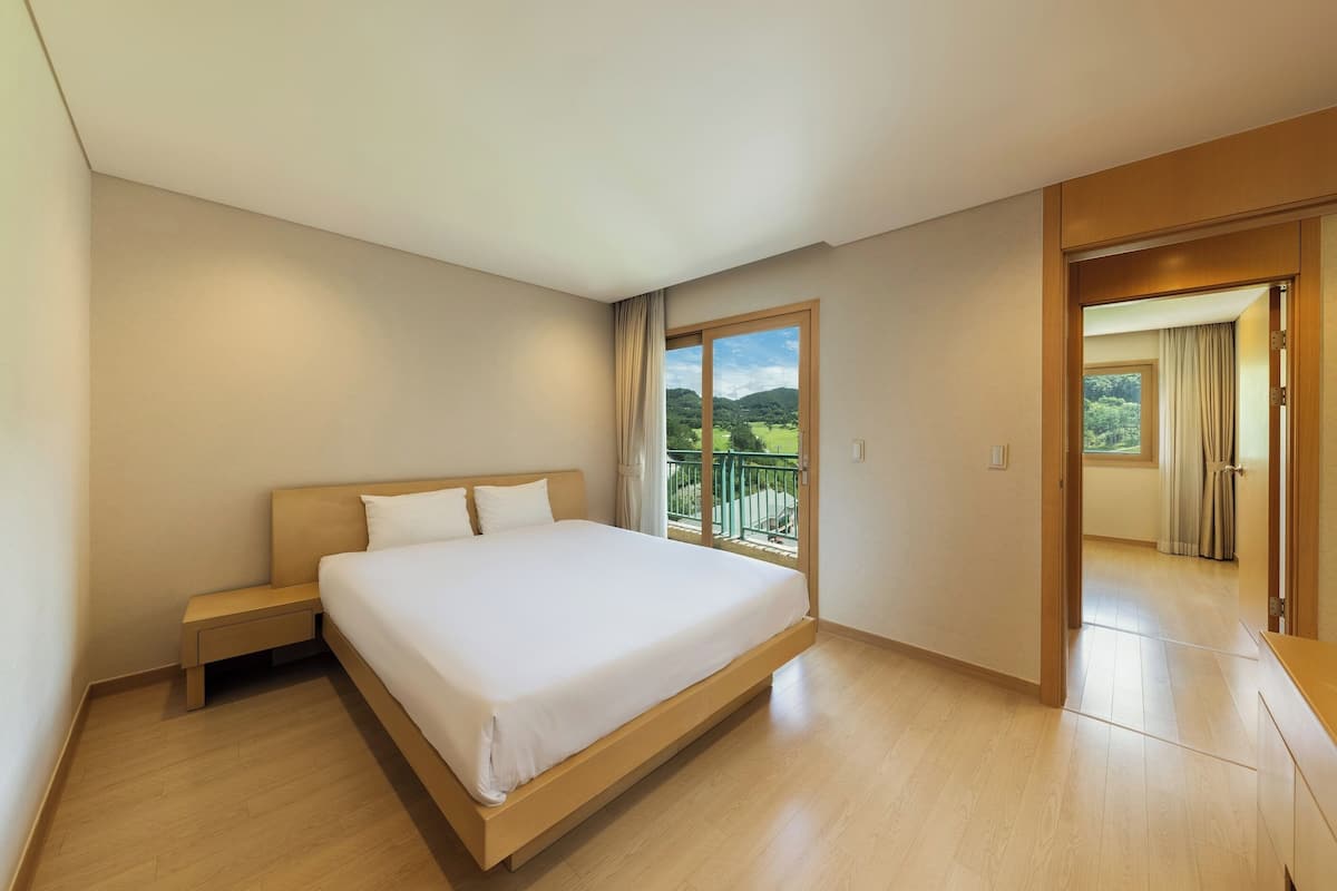 A bedroom with a large bed, a nightstand, a window with a view of greenery, and a door leading to another room.