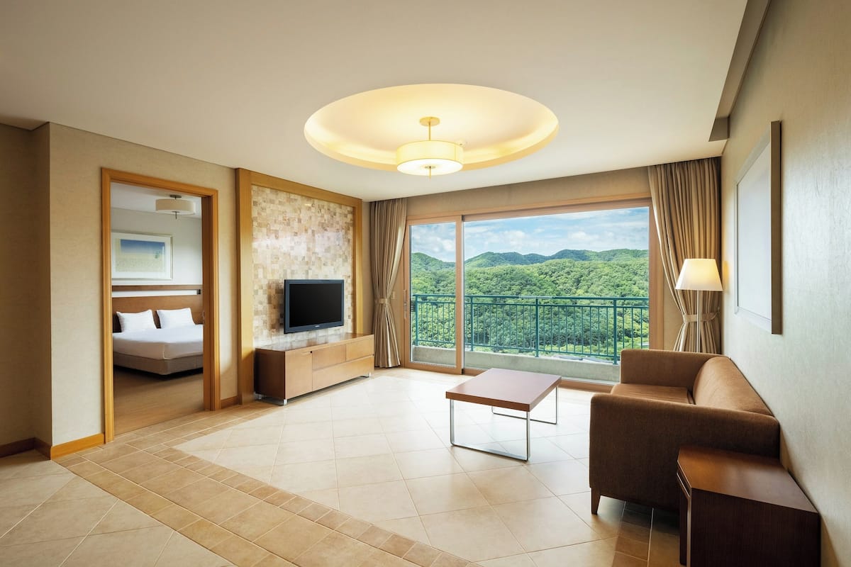 A hotel room with a bed, a TV, a sofa, a table, and a balcony with a view of the mountains.