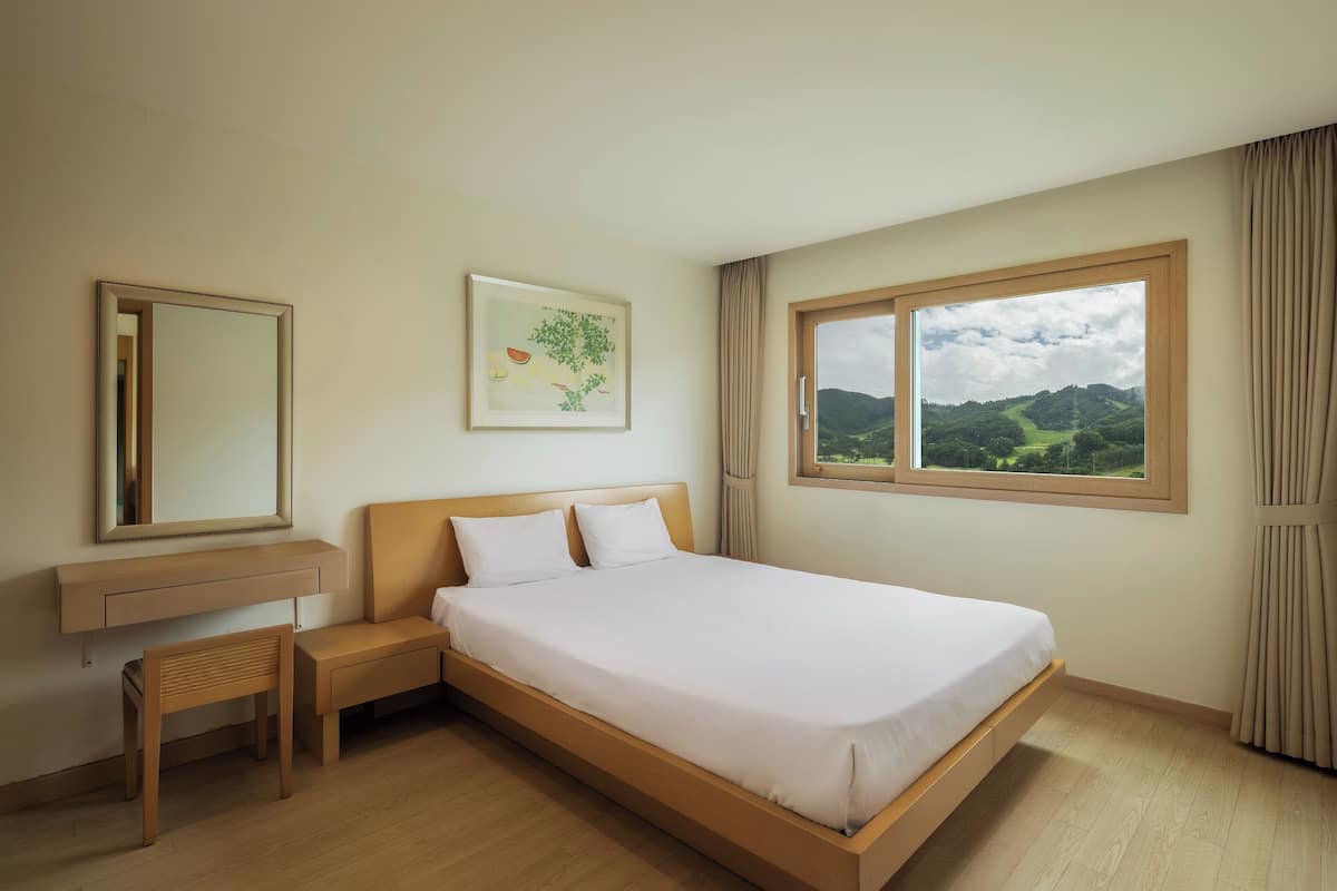 A hotel room with a bed, a bedside table, a mirror, a window with a view of hills, and a painting on the wall.