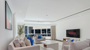 Ein modernes Wohnzimmer mit einer grauen Sofagarnitur, einem Couchtisch und einem Flachbildfernseher, der 'NETFLIX' anzeigt. Der Raum ist minimalistisch eingerichtet, mit weißen Wänden und einem großen Fenster, das für natürliches Licht sorgt.