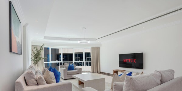 A modern living room with a grey sofa set, a coffee table, and a flat-screen TV displaying 'NETFLIX'. The room has a minimalist design with white walls and a large window providing natural light. - LUX The Al Seef Marina Suite 2 (Dubai)