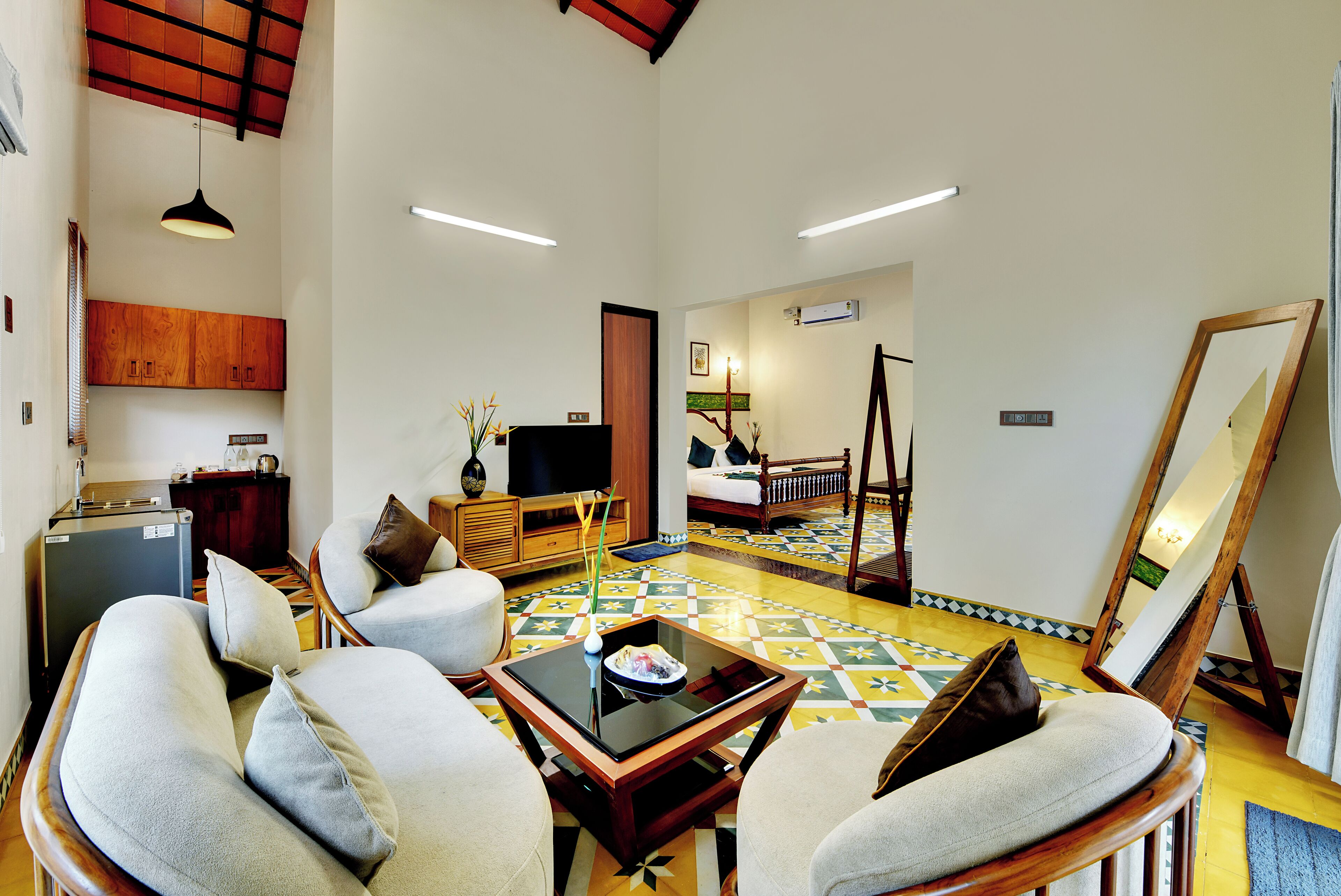 Amplia sala de estar con un conjunto de sofás, una mesa de centro y una mesa de comedor. Se aprecia una pequeña cocina con refrigerador y armarios. En las paredes se ven un espejo grande y un cuadro.