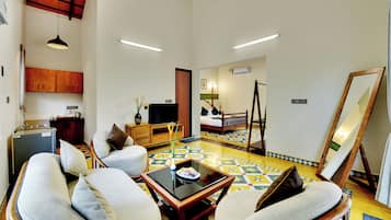 Amplia sala de estar con un conjunto de sofás, una mesa de centro y una mesa de comedor. Se aprecia una pequeña cocina con refrigerador y armarios. En las paredes se ven un espejo grande y un cuadro.