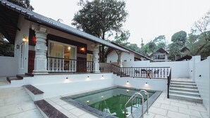 Ein Poolbereich mit Glasboden, Stufen zu einer überdachten Terrasse und ein Gebäude mit Säulen und einem Balkon.