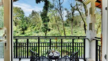 A balcony with a view of a lush forest, two chairs, a small table with a teapot, and a decorative candle.
