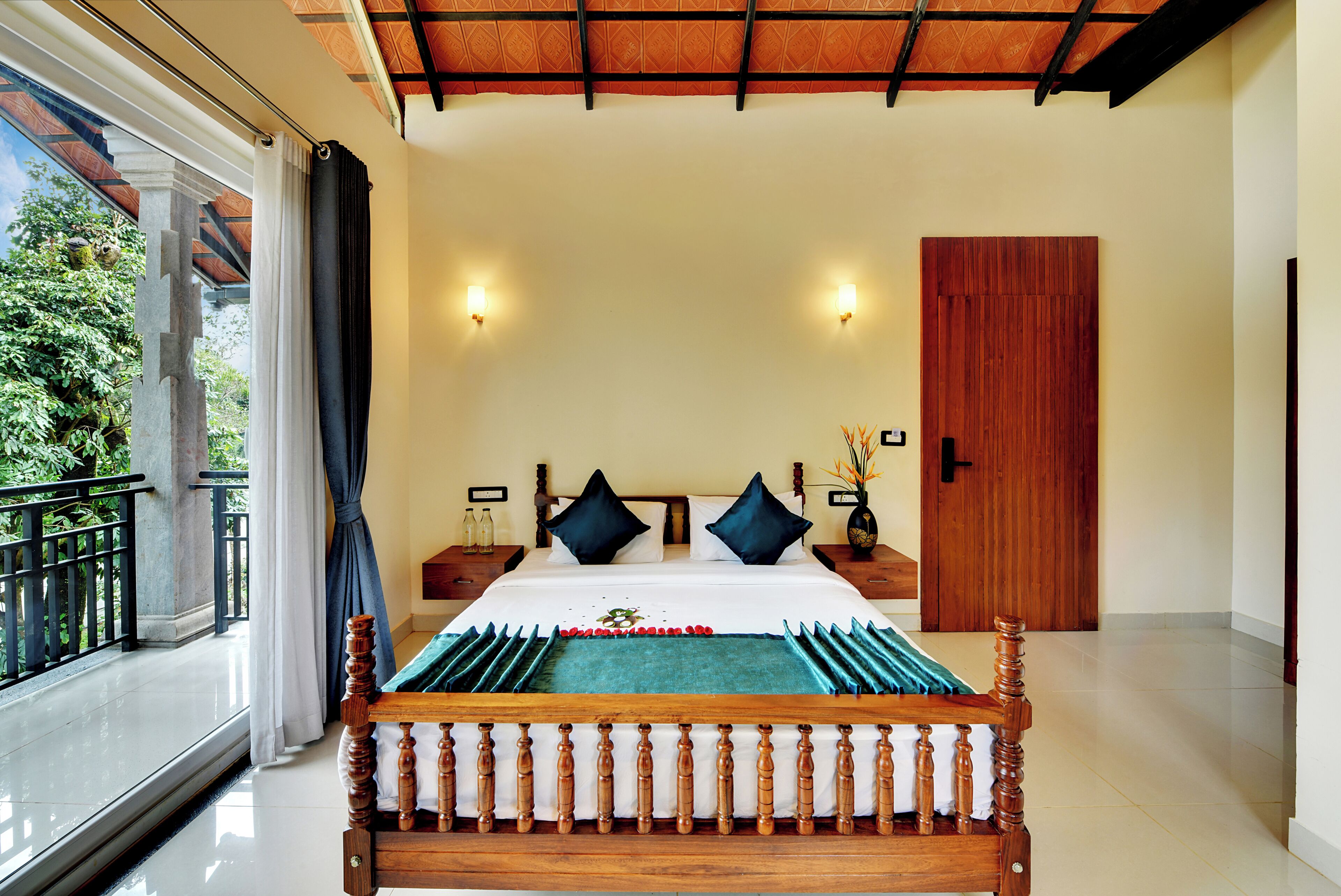 A bedroom with a large bed, two nightstands, a wooden door, and a view of greenery through a balcony.