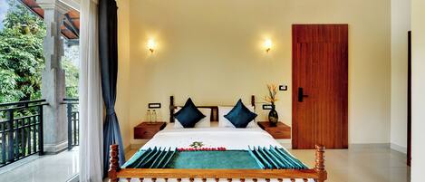 A bedroom with a large bed, two nightstands, a wooden door, and a view of greenery through a balcony.