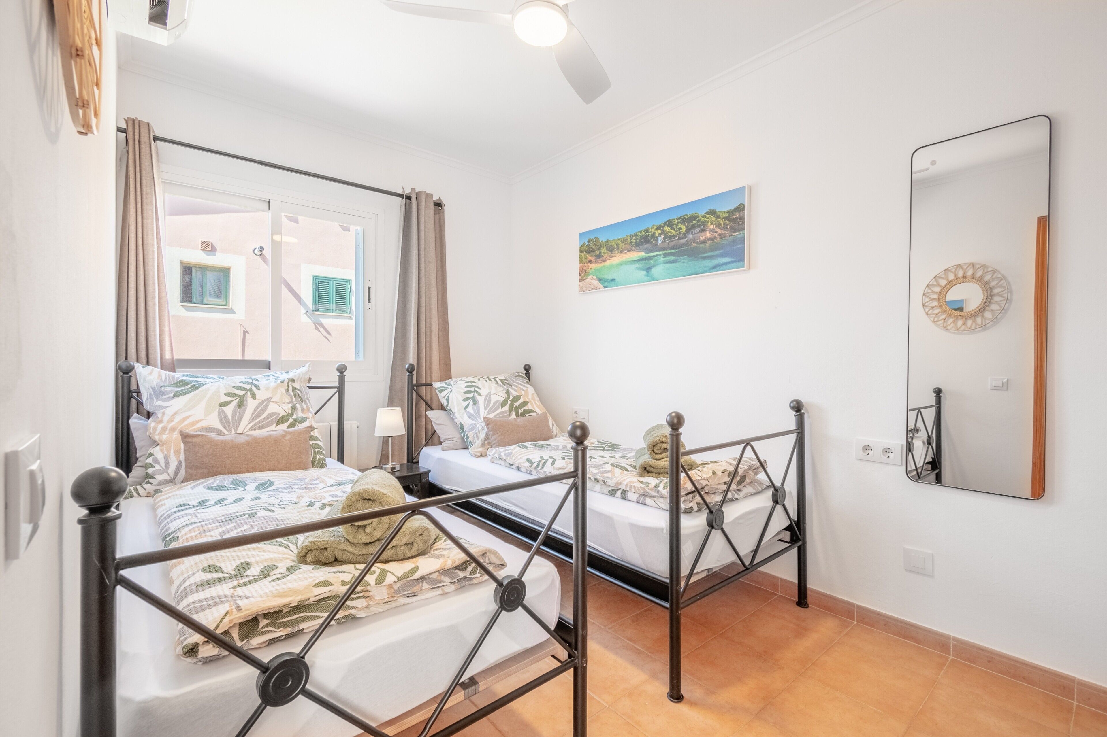 Een slaapkamer met twee bedden, een plafondventilator, een schilderij, een spiegel en uitzicht op een gebouw.