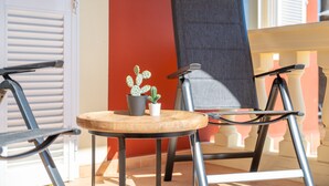 Una silla con estructura metálica y asiento de madera, una pequeña mesa redonda con una planta en maceta, y una ventana con contraventanas blancas.