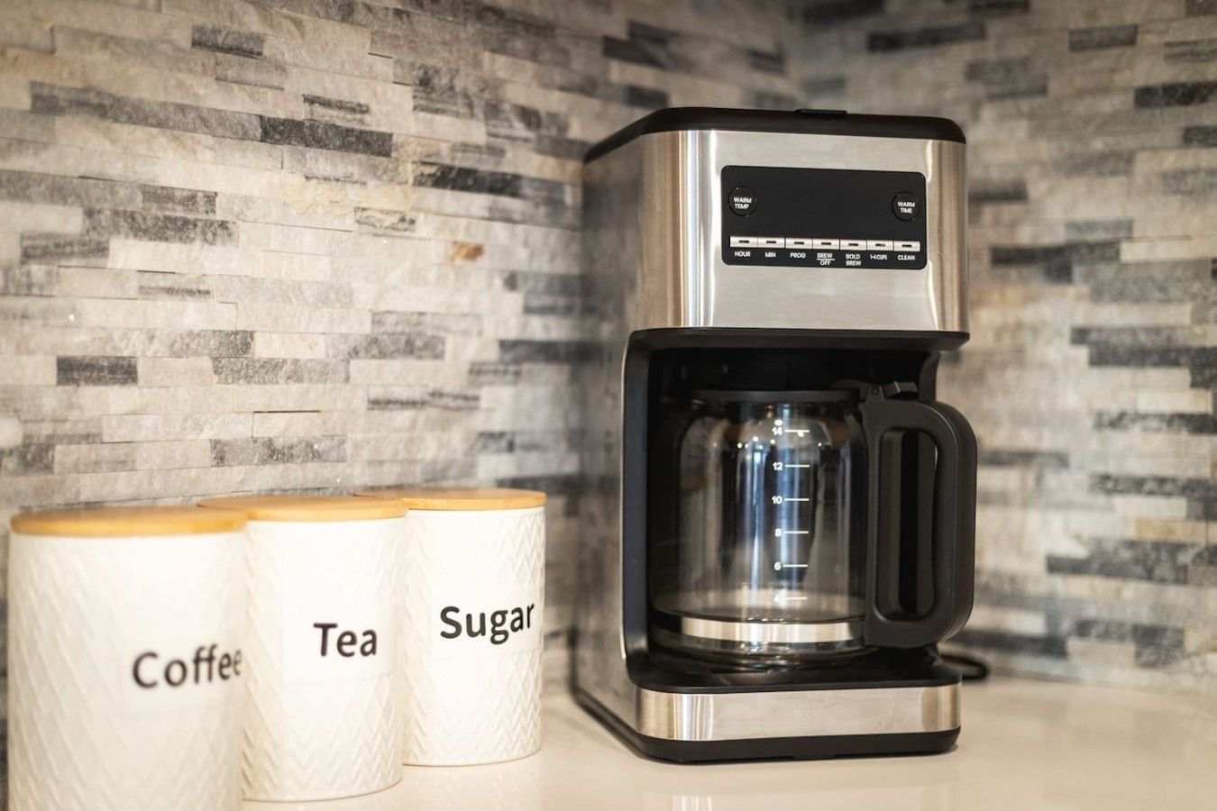 A coffee maker with a digital display, next to containers labeled 'Coffee,' 'Tea,' and 'Sugar.'