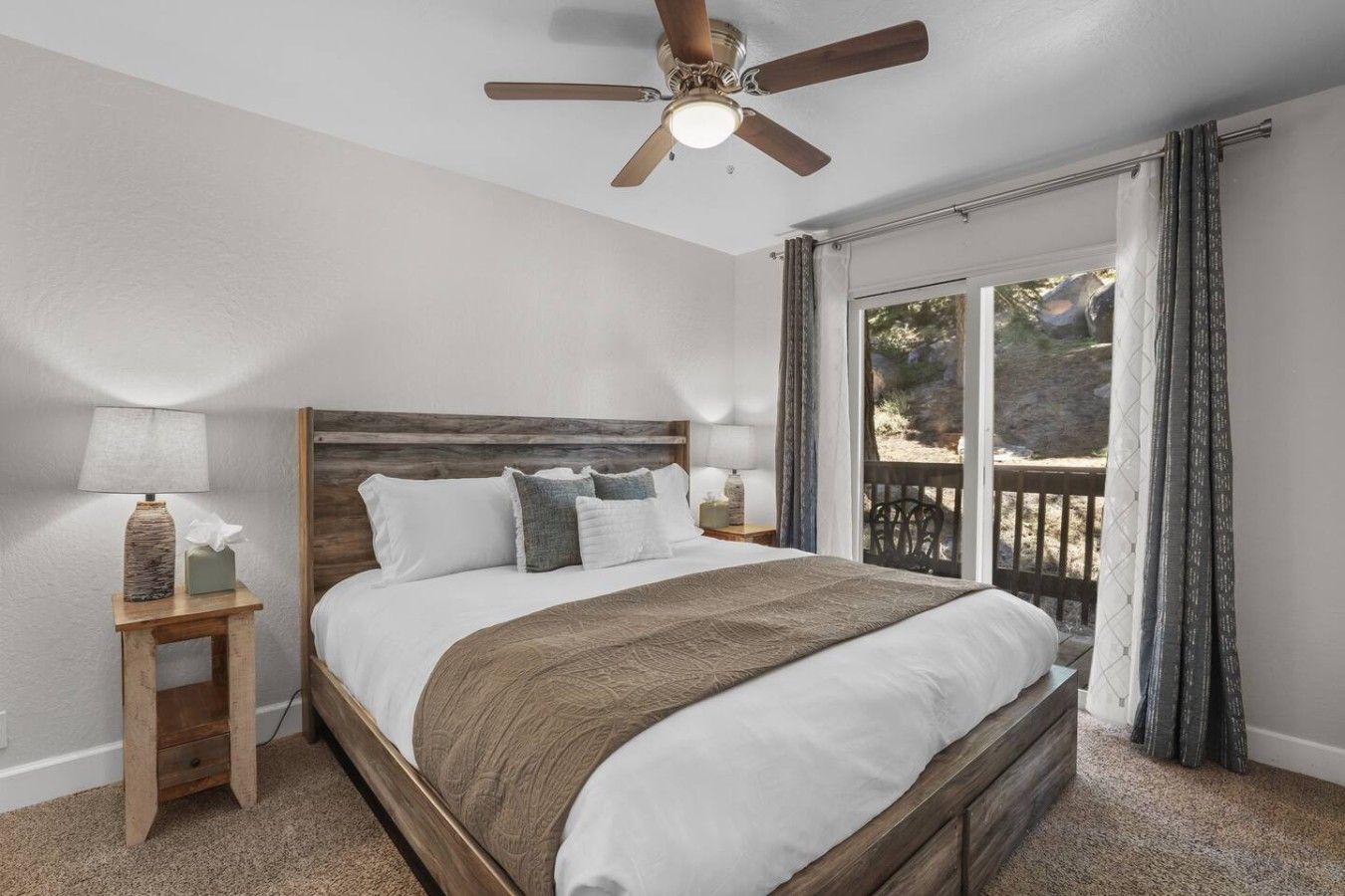 Quarto com cama de madeira, duas mesas de cabeceira com luminárias, um ventilador de teto e vista para uma varanda.