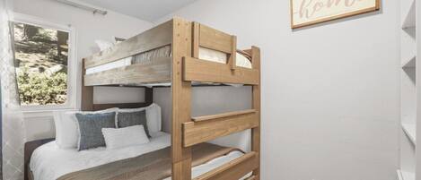 Una cama litera con estructura de madera, ropa de cama blanca y una ventana con vistas a zonas verdes.
