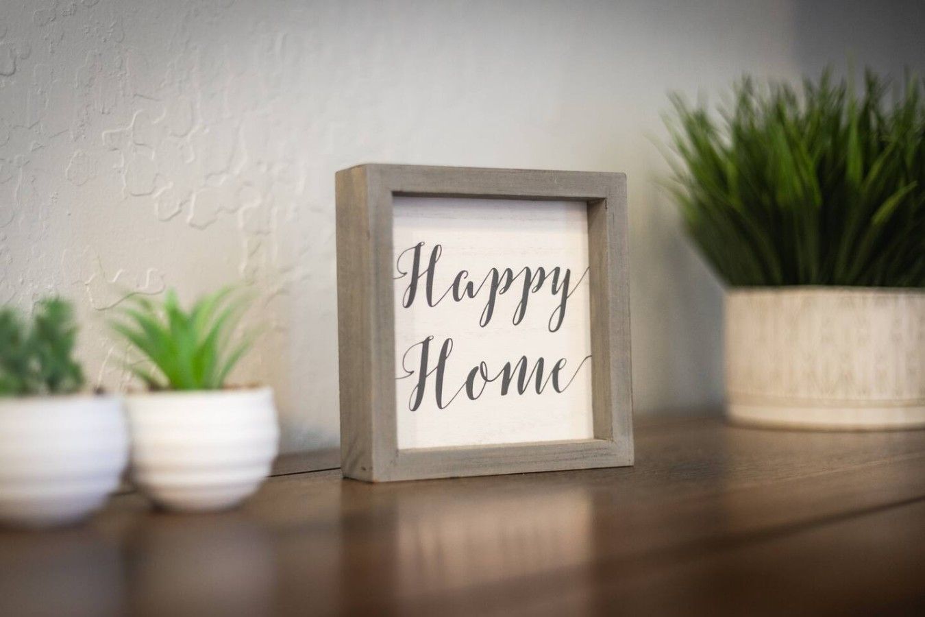 Ein gerahmtes Schild mit dem Schriftzug 'Happy Home' auf einer Holztischplatte, begleitet von zwei Topfpflanzen.