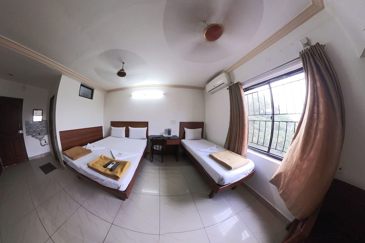 Habitación de hotel con dos camas individuales, un escritorio, una silla, una ventana con cortinas, un ventilador de techo y un baño con lavabo y espejo.