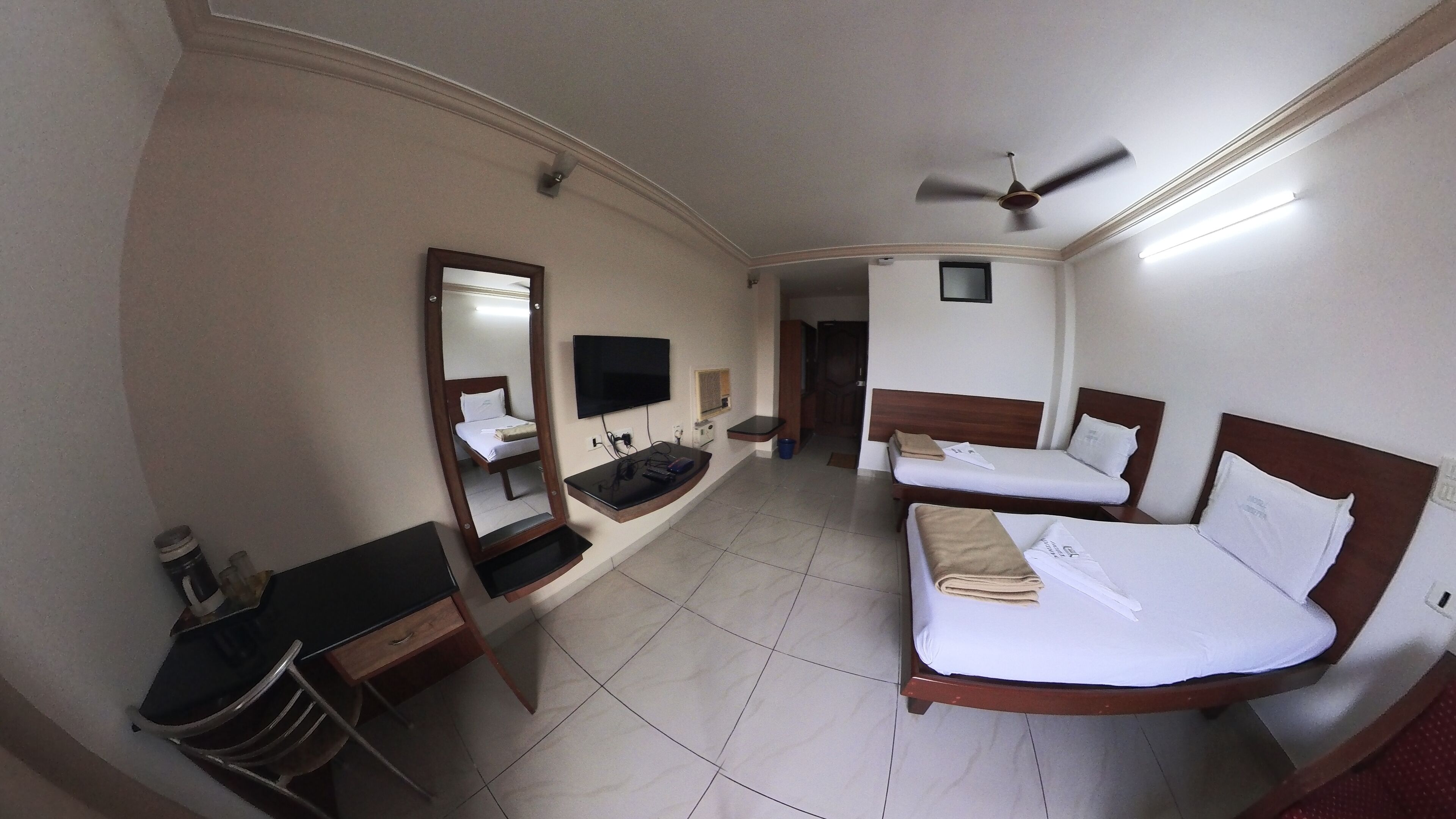 Ein Hotelzimmer mit Bett, Schreibtisch, Stuhl, Spiegel, Fernseher und Deckenventilator.